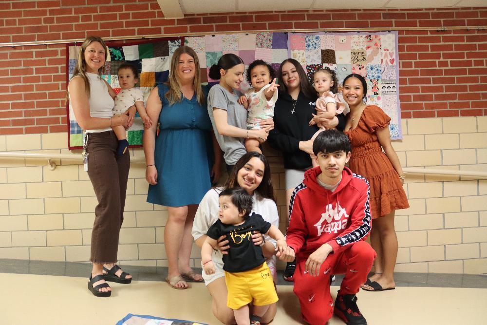 A group of adults and toddlers pose for a photo in front of a wall-hanging quilt. The adults hold and surround the children. The background is a brick wall.