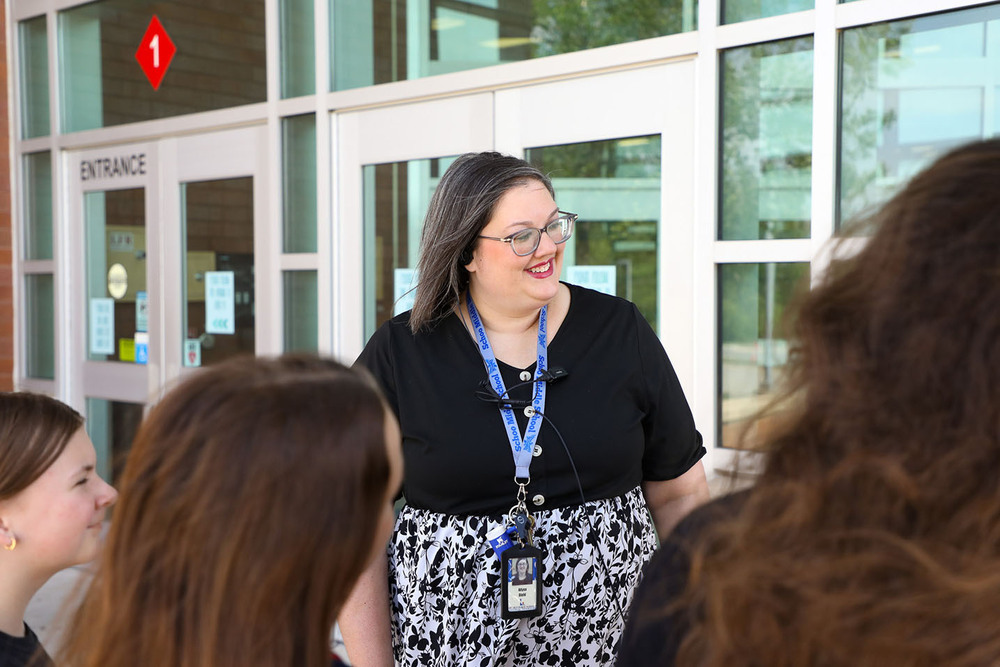 Schoo Middle School Principal Alyssa Diehl stands outside the main entrance of the school building. She is smiling while talking with three girls who are in front of her. There are windows and doors behind all of them.