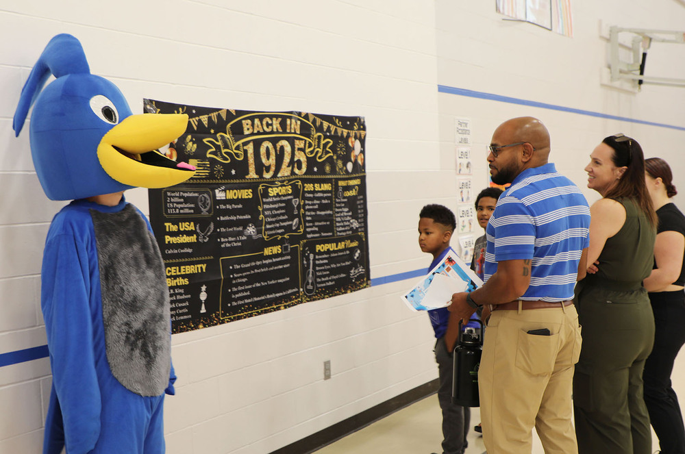 Randolph Elementary School community members look at a poster that says Back in 1925 during a centennial celebration at the school building. A person dressed up in a Roadrunner costume is at the lefthand side of the photo.
