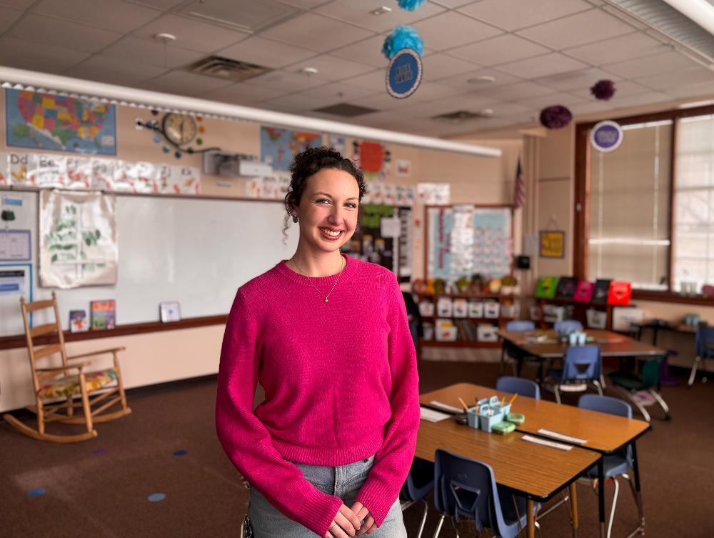 A smiling woman stands in a brightly lit elementary classroom, wearing a pink sweater and jeans.