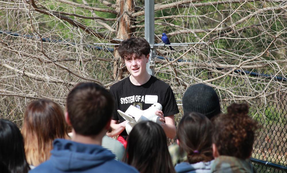 A young man presents to an outdoor group, holding a white object. A bright blue bird perches on a metal pole behind him. The background is a dense thicket of bare branches.