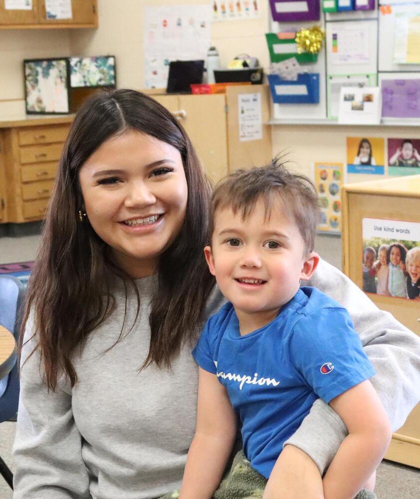 A young woman smiles while holding a toddler on her lap in a classroom. The toddler wears a blue shirt that says "Champion." "Use kind words" is visible on a nearby poster.