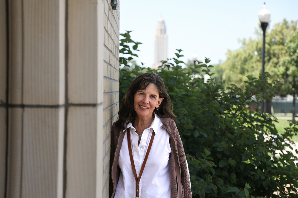 A smiling woman leans against a brick building outdoors with the Nebraska State Capitol blurred in the background.