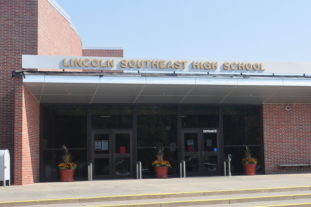 The front entrance of Lincoln Southeast High School is shown. There are two sets of doors and windows underneath a metal awning with the words 'Lincoln Southeast High School' on it.