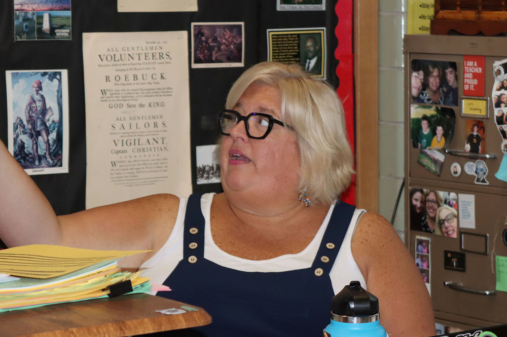 Lefler Middle School social studies teacher Becky Boswell points to a screen while sitting at her classroom desk. She is facing right to left and is looking at the screen. Multiple historical documents and photos are attached to a bulletin board behind her.
