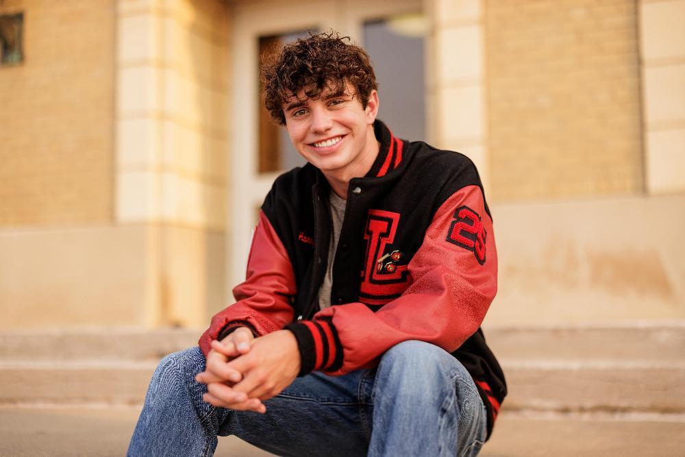 A young man, wearing a red and black letterman jacket, sits on steps outside a building, smiling at the camera. The jacket has "L" and "23" embroidered on it.