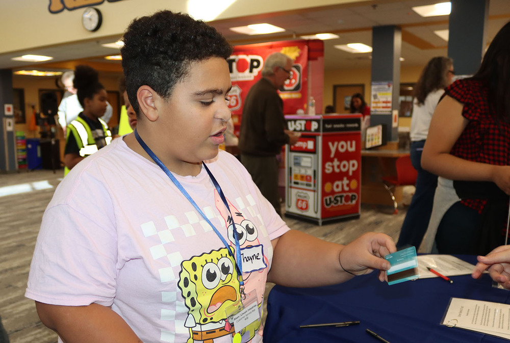 Culler Middle School sixth grader Thayne receives a debit card from a bank during the BizTown simulation activity. He is facing left to right and is taking the debit card with his left hand. He is standing in front of other people in the Culler library.