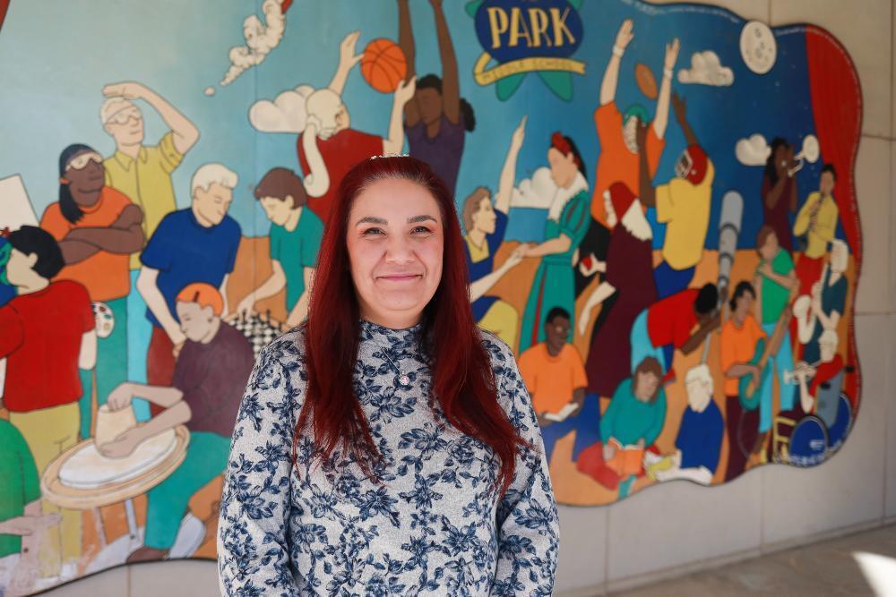 A smiling woman with long reddish-brown hair stands before a vibrant mural. It depicts diverse cartoon-style people engaging in activities like sports, music, art, and chess, with "PARK MIDDLE SCHOOL" at the top.