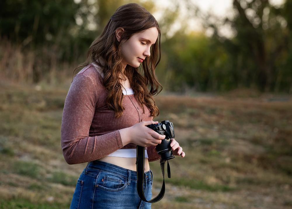 A young woman holds and examines a DSLR camera outdoors. She's wearing a mauve sweater and jeans in a natural, grassy setting.