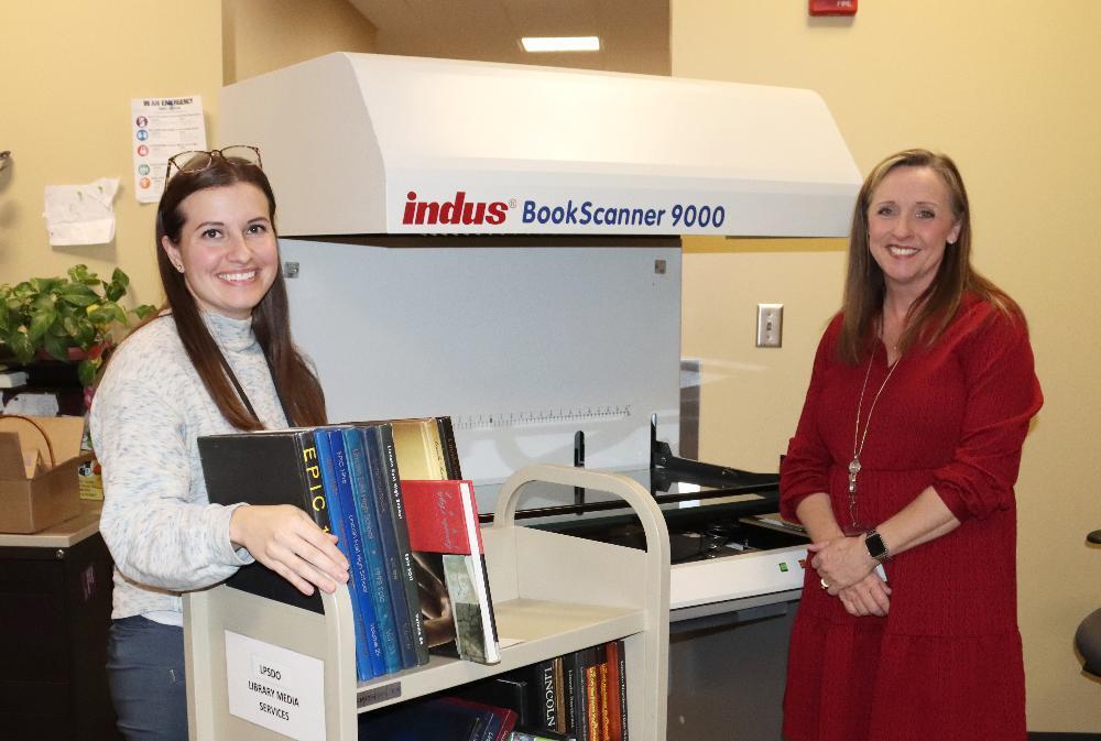 From left, Sara Scott and Chris Haeffner smile next to an Indus BookScanner 9000 machine. They use the machine to scan yearbook pages for a digitization project. Sara is at the left of the picture and has long hair and is wearing glasses on her forehead. Chris is at the right and has shoulder-length hair. Sara's right hand is by a library cart full of yearbooks that are waiting to be scanned for the project.