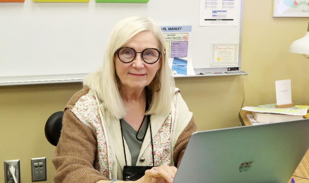 Judy Wadhams smiles while sitting at her desk in a classroom at Schoo Middle School. Her hands are over the keyboard of her laptop computer and the back of the screen is facing the camera. She has shoulder-length hair and is wearing glasses and an audio device to project her voice to students.
