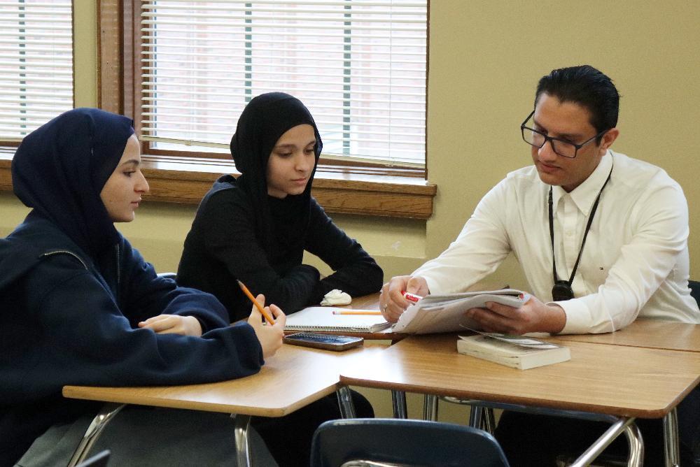 Three students, two women wearing hijabs and one man, sit at desks in a classroom. They review papers together, engaging in a study session.