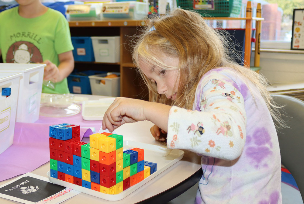 Holmes Elementary School first grader Marceline carefully places a block on top of a castle she is making on a classroom table. She is reaching her left hand over the tower to made adjustments to the block. She is facing right to left.