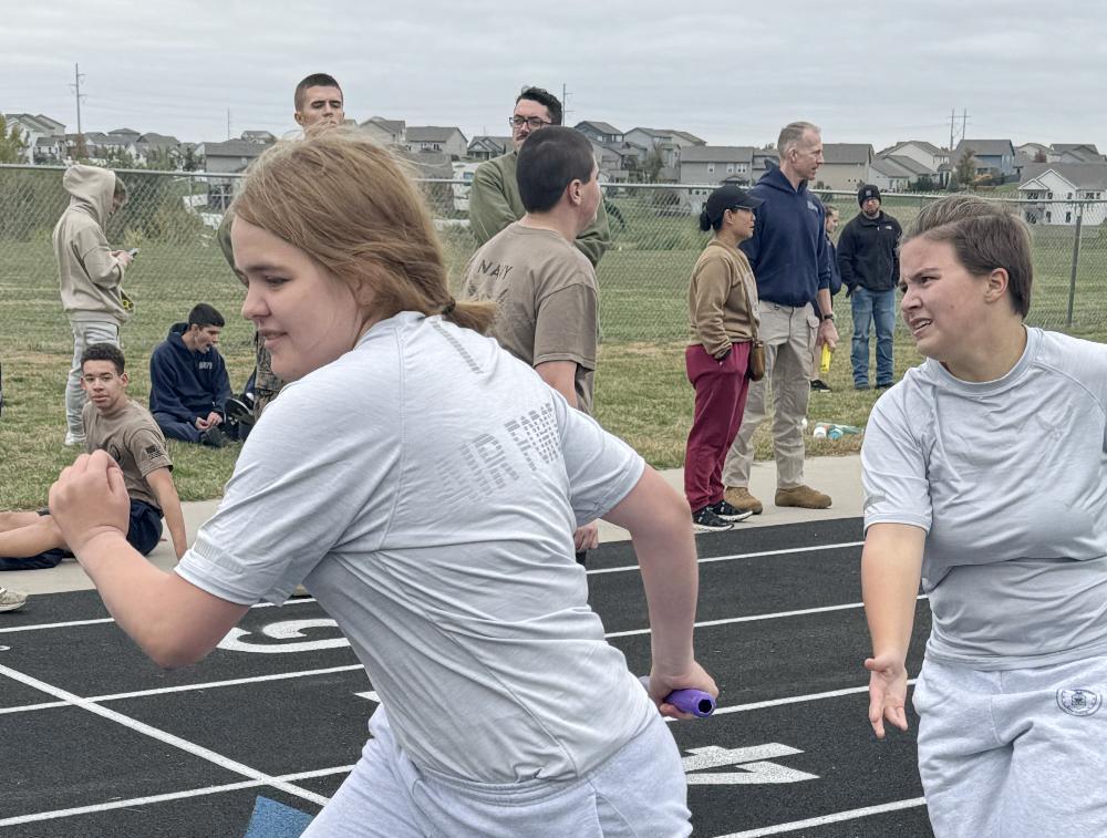 Two girls are running in an outdoor relay race on the track. One girl has just handed the baton to her teammate with her right hand at the far righthand side of the photo. A girl in the middle of the picture has taken the baton with her right hand and is preparing to sprint away down the track. Several students are watching the race from the edge of the track.