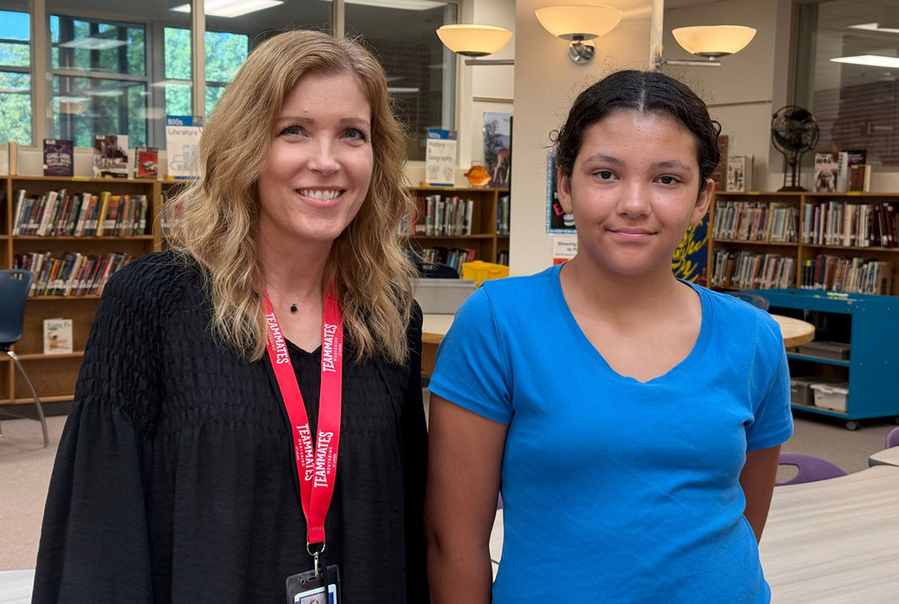 From left, TeamMates mentor Stacey and mentee Aivah stand together in a school library. There are tables and shelves full of books and library materials in the background.