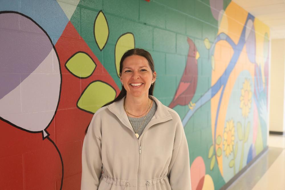 A happy woman in a light jacket smiles, centered against a colorful brick wall mural. The mural depicts abstract shapes, a large balloon, a red bird, and yellow flowers.