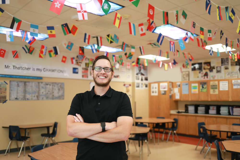 A smiling teacher wearing glasses and a black polo shirt stands with his arms crossed in a classroom decorated with international flags hanging from the ceiling. Desks and educational materials are visible in the background.