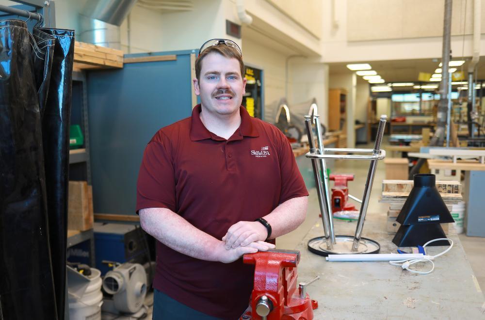 A person wearing a maroon polo shirt and safety glasses stands in a workshop, resting their hands on a red vise mounted to a workbench, with tools and metal equipment visible in the background.