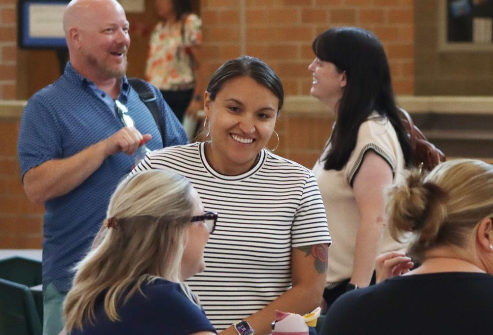 A Lincoln Public Schools leader smiles as she stands next to a table and speaks to another person who is sitting at the table. Two other people are smiling in the background as they are having a separate conversation.