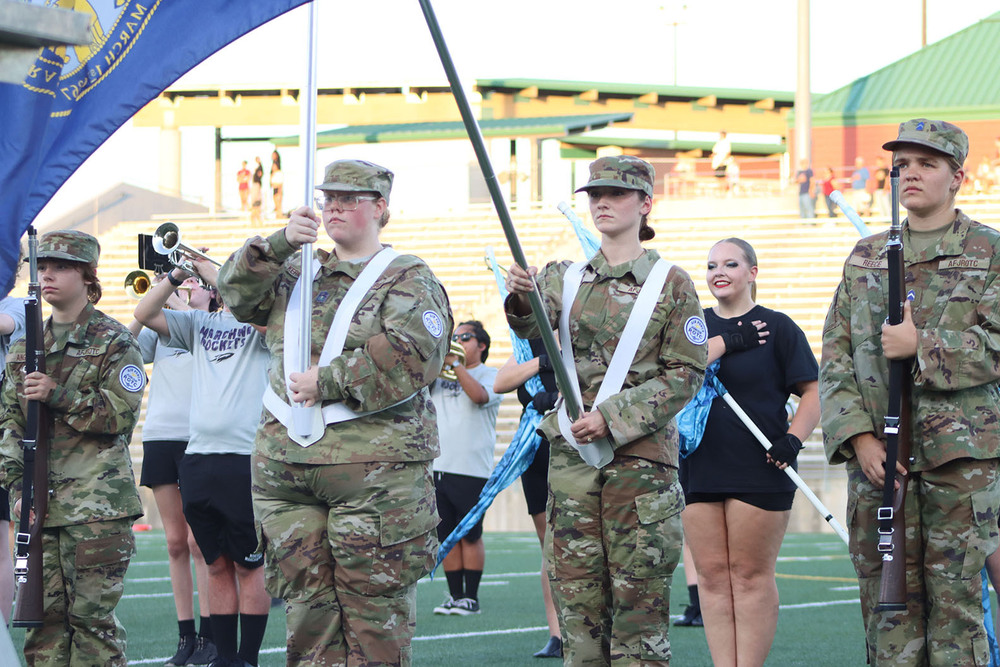 From left, Air Force Junior Reserve Officer Training Corps cadets Ally, Gabrielle, Trinity and Kriss stand in a row at midfield of Seacrest Field for their color guard presentation. Ally and Kriss are both carrying ceremonial rifles, and Gabrielle and Trinity are holding poles attached to the Nebraska state flag and the United States flag. Northeast High School marching band members are in the background.