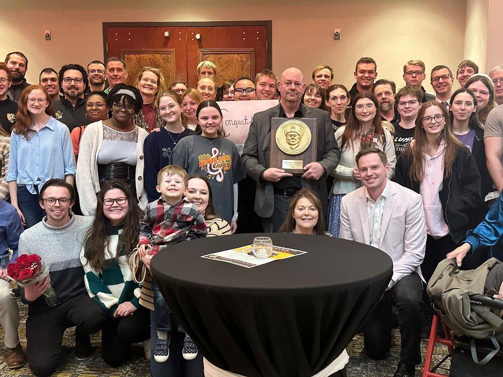 R.J. Metteer holds a plaque for his state band award. He is surrounded by more than two dozen supporters he met throughout his time at Southeast High School. They are standing and kneeling in four rows behind a round table.