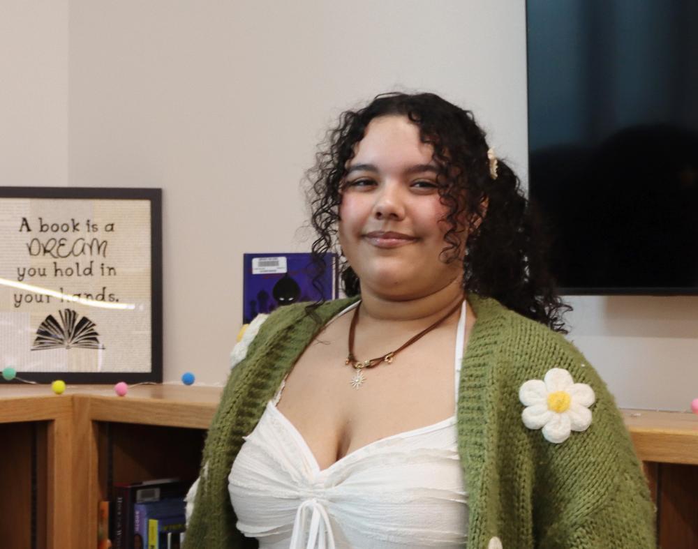 A young woman with curly hair is posing in a library. She wears a green cardigan with flower accents and a white top. A framed quote reads: "A book is a dream you hold in your hands."