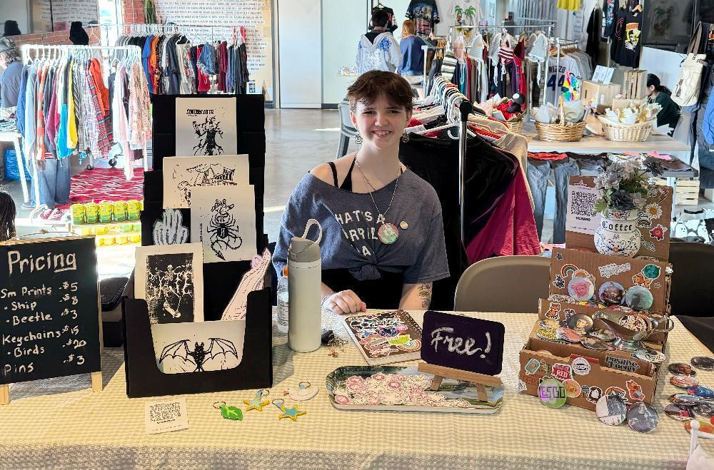 A young woman sits at a table, displaying and selling small prints, keychains, pins, and stickers at an indoor market. Pricing is shown on a nearby chalkboard.