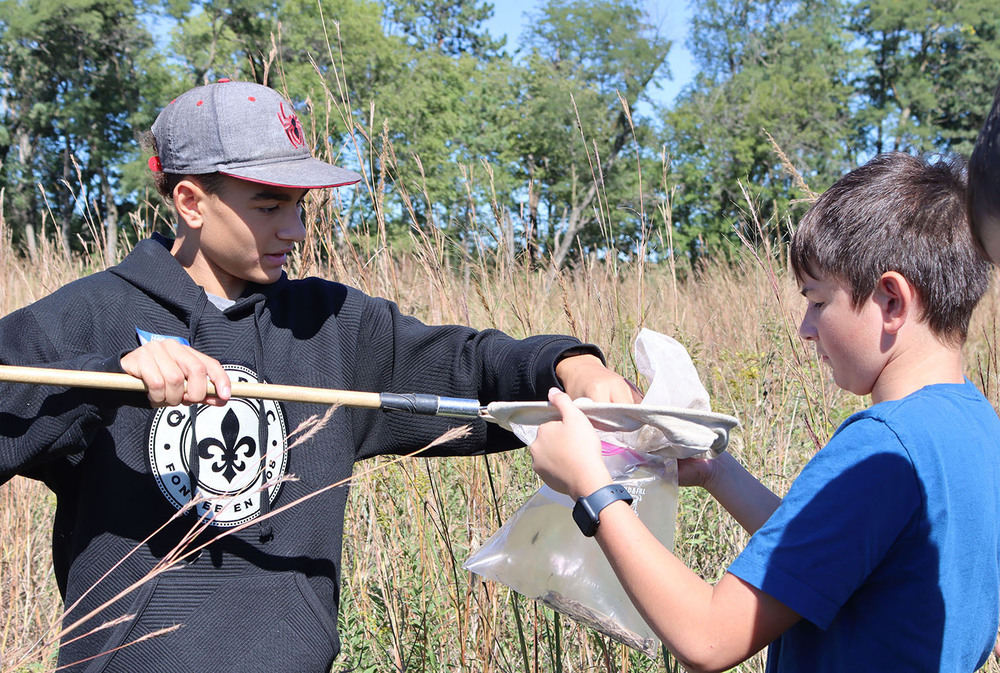 From left, Lefler Middle School seventh graders Uriah and Cooper work on collecting insects and arachnids while standing in a tallgrass prairie. Uriah is emptying a large bug net into a plastic collection bag that Cooper is holding with both hands.