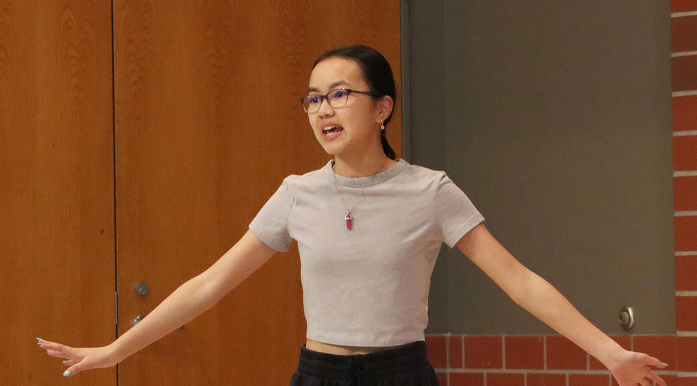 A Scott Middle School student performs on stage during a visit by two professional actors. She is extending both hands away from her body.
