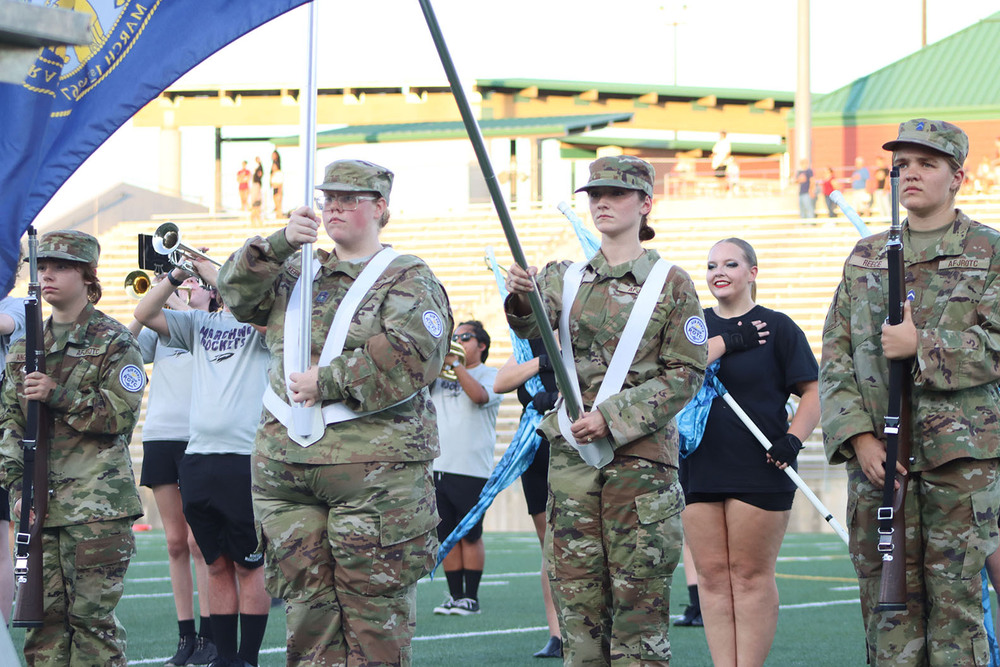 From left, Air Force Junior Reserve Officer Training Corps cadets Ally, Gabrielle, Trinity and Kriss stand in a row at midfield of Seacrest Field for their color guard presentation. Ally and Kriss are both carrying ceremonial rifles, and Gabrielle and Trinity are holding poles attached to the Nebraska state flag and the United States flag. Northeast High School marching band members are in the background.