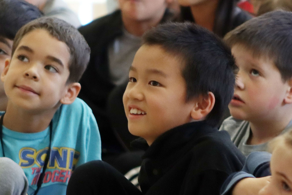 Three Kooser Elementary School students smile as they watch UNL student-athletes speak during a presentation.