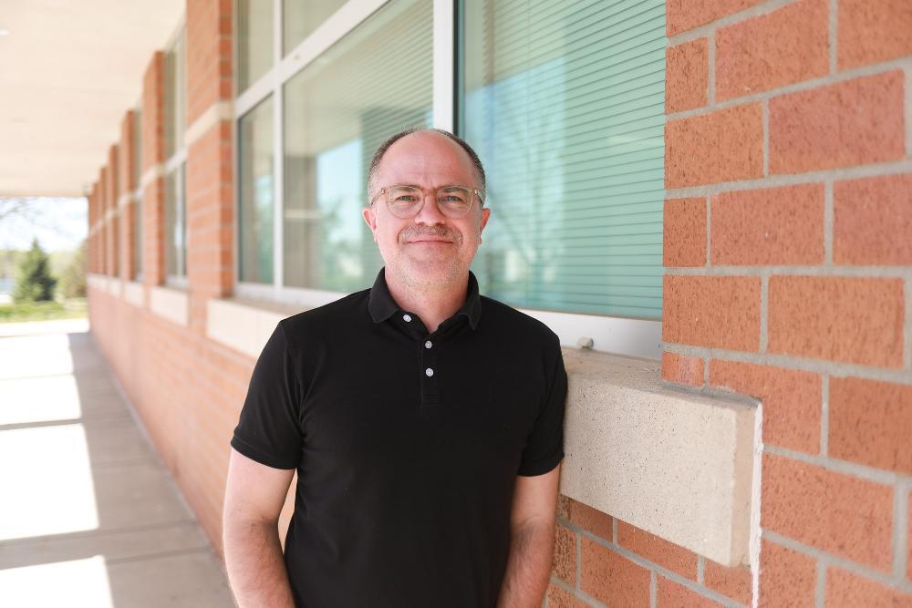 A smiling, bald, middle-aged man with glasses and a mustache wears a black polo shirt. He stands outdoors, leaning against a brick building with large windows and horizontal blinds. Bright sunlight illuminates the scene.