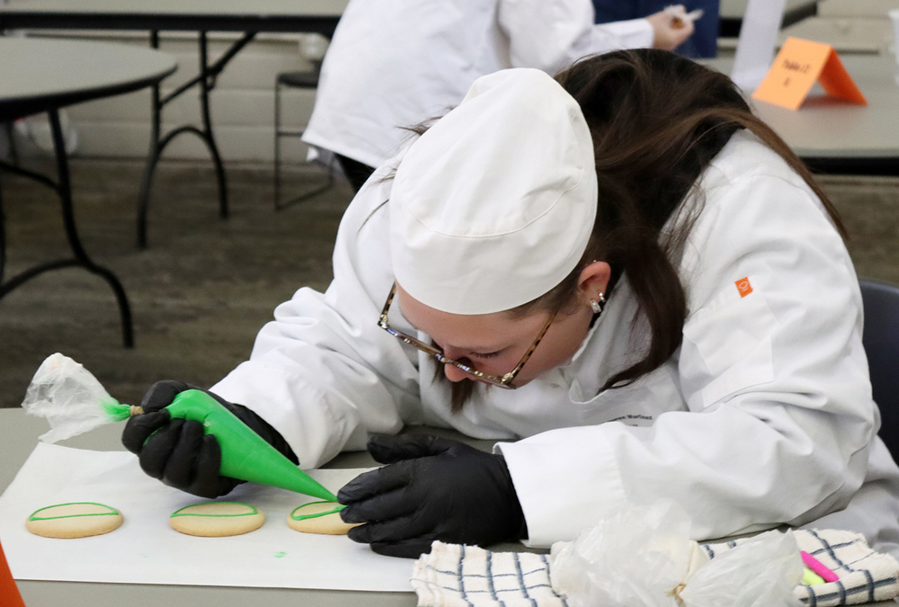 A student carefully places frosting on a row of cookies by squeezing a tube at the LPS Culinary Contest. She is facing right to left and is bending down over one of the cookies on a table.