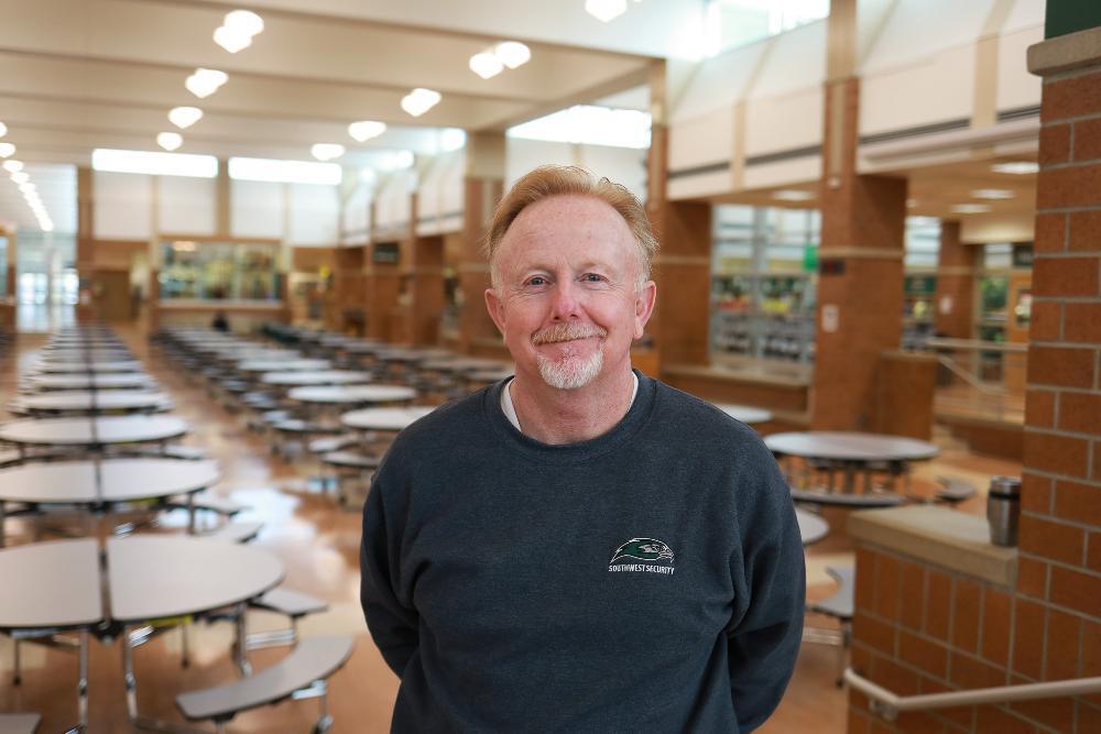 A smiling man with reddish-blonde hair and a goatee stands in a large, bright, empty cafeteria. He wears a dark gray sweatshirt with a "Southwest Security" logo. Rows of round tables with attached seats fill the background.
