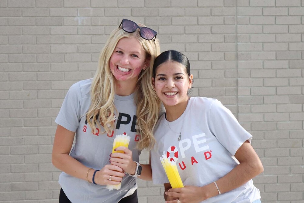 Two North Star High School students stand in front of a brick wall at Union Bank Stadium. They are wearing t-shirts that say Hope Squad on them and are preparing to hand out glow sticks to fans at a football game.