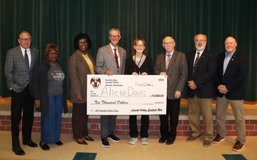 Eight people stand smiling. Central woman, Alicia Davis, holds a giant $10,000 check from Scottish Rite Lincoln Valley. She's recognized as "LPS Teacher of the Year." They are in front of a green curtain and brick wall.