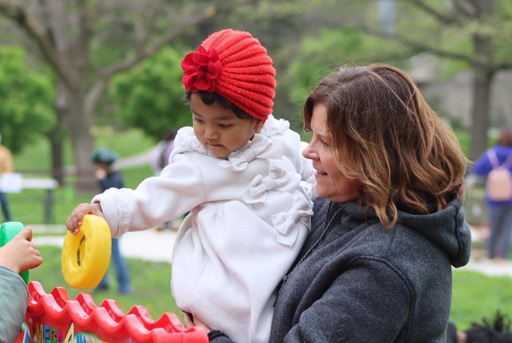 A toddler, wearing a red hat and white coat, manipulates a yellow toy at a playground; a woman holds the toddler.