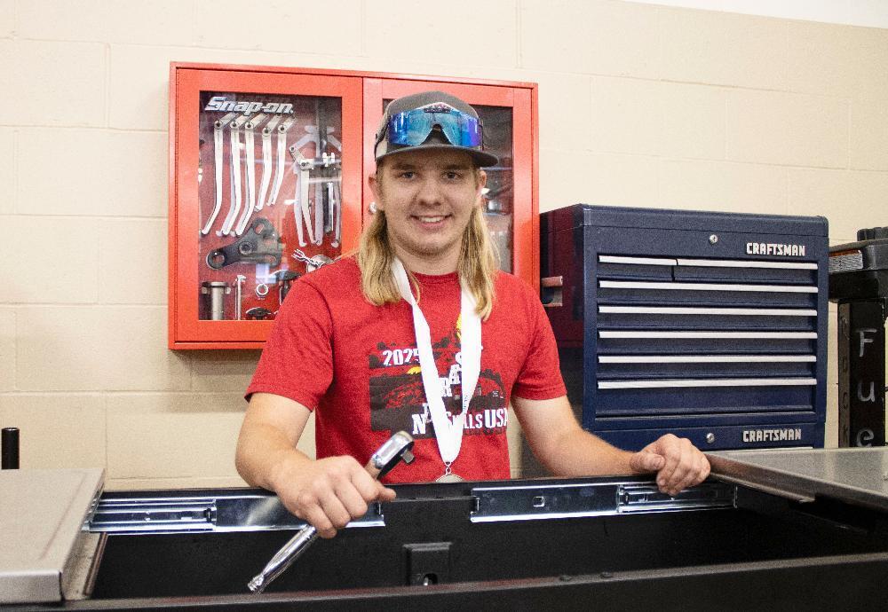 Northeast High School graduate Jaxson stands in front of a glass case full of small silver tools and behind a toolbox full of assorted hand tools. He is wearing a ballcap with sunglasses on the bill of the cap and is smiling while holding a tool in his right hand.