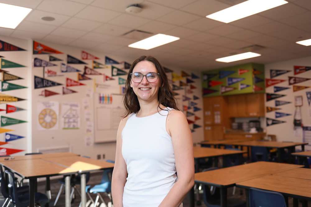 A woman wearing glasses and a white sleeveless top smiles in a classroom with tables, chairs and college pennants on the walls.