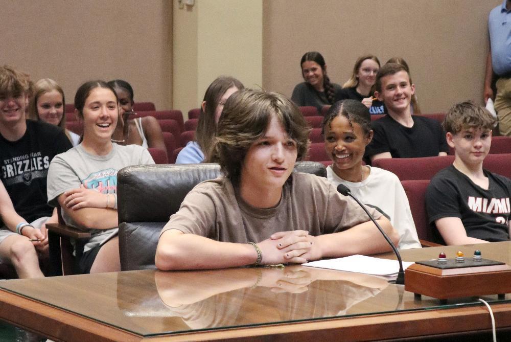 A young person sits at a table, speaking, in an assembly hall. Other young people and adults are seated around.