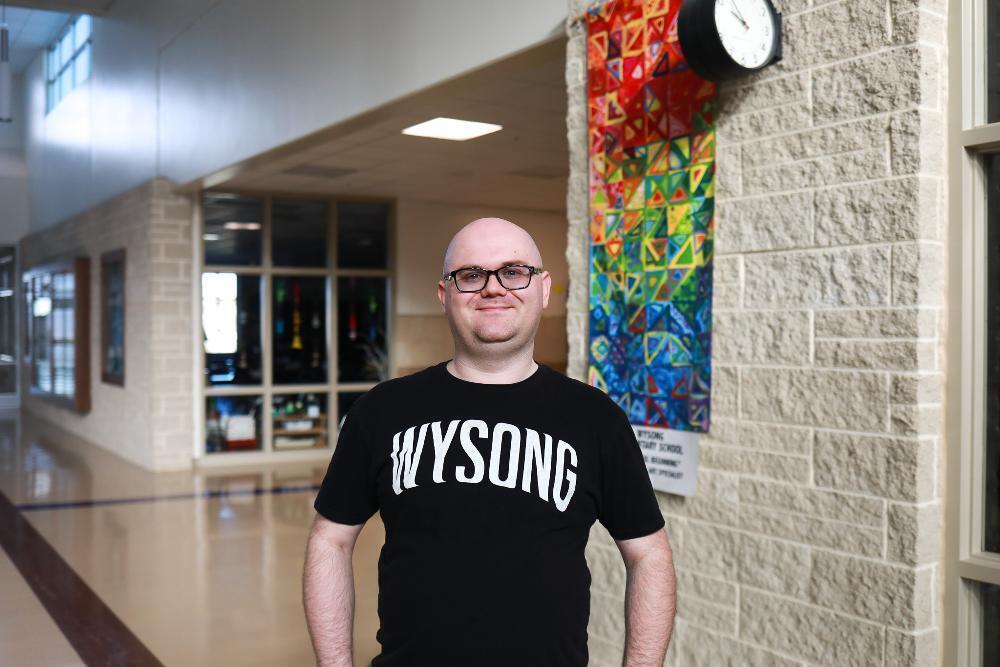 A smiling bald man with glasses in a black "WYSONG" t-shirt stands in a school hallway. Behind him, a colorful quilt and clock hang on a stone wall, with a long corridor extending left.