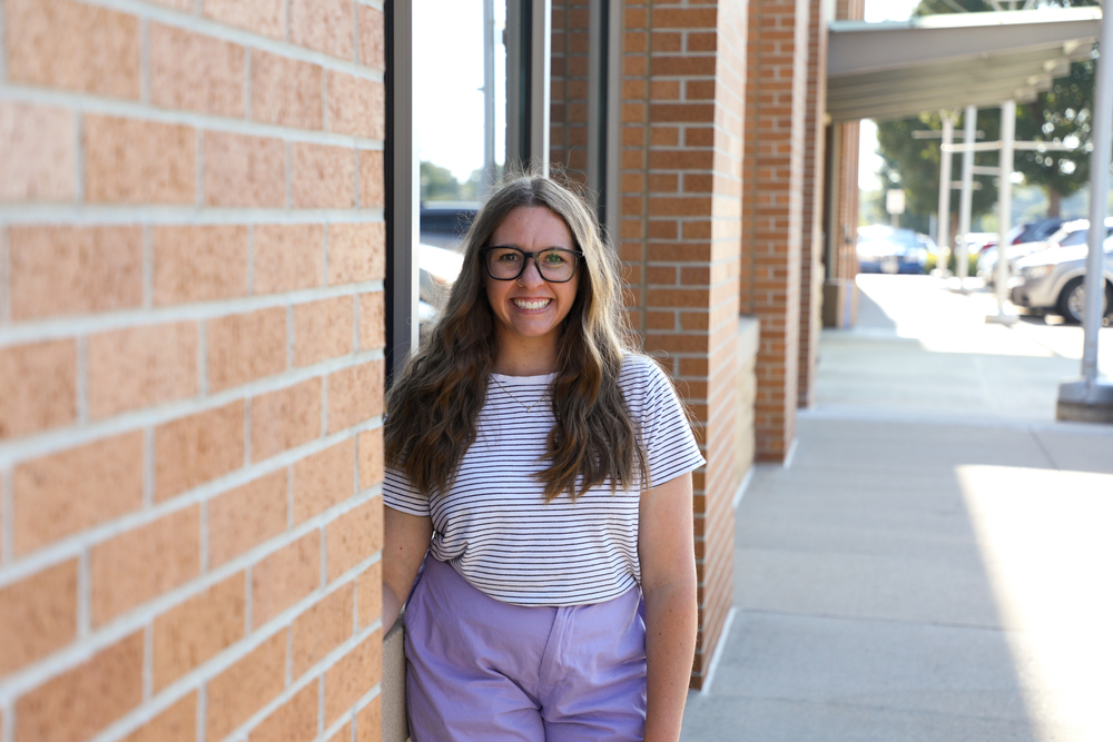 A woman with brown hair and glasses wearing a striped shirt and purple pants stands next to a brick building