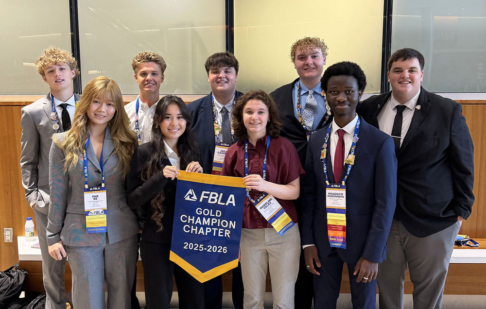 Nine Southeast High School students are standing in front of a wall panel and windows at the FBLA National Leadership Conference. The three girls and six boys are holding a banner that says 'FBLA Gold Champion Chapter 2025-2026' on it.