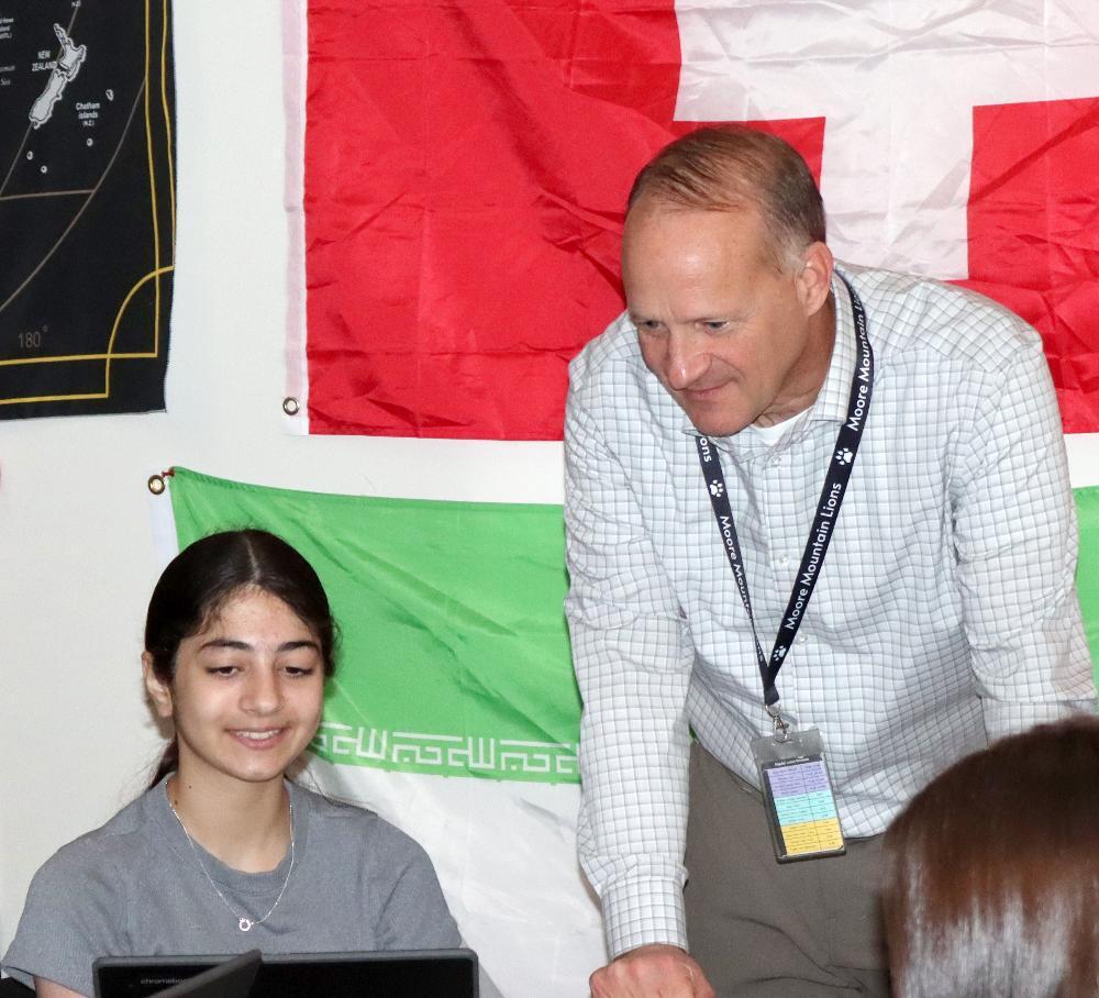 From left, a student smiles with Moore Middle School Principal Gary Czapla in a social studies classroom. The student is sitting at a desk with her laptop in front of her on the lefthand side of the photo. Gary is on the righthand side and is slightly bending over at the table to see what is on her laptop screen. There are flags of different world countries pinned on the wall behind them.