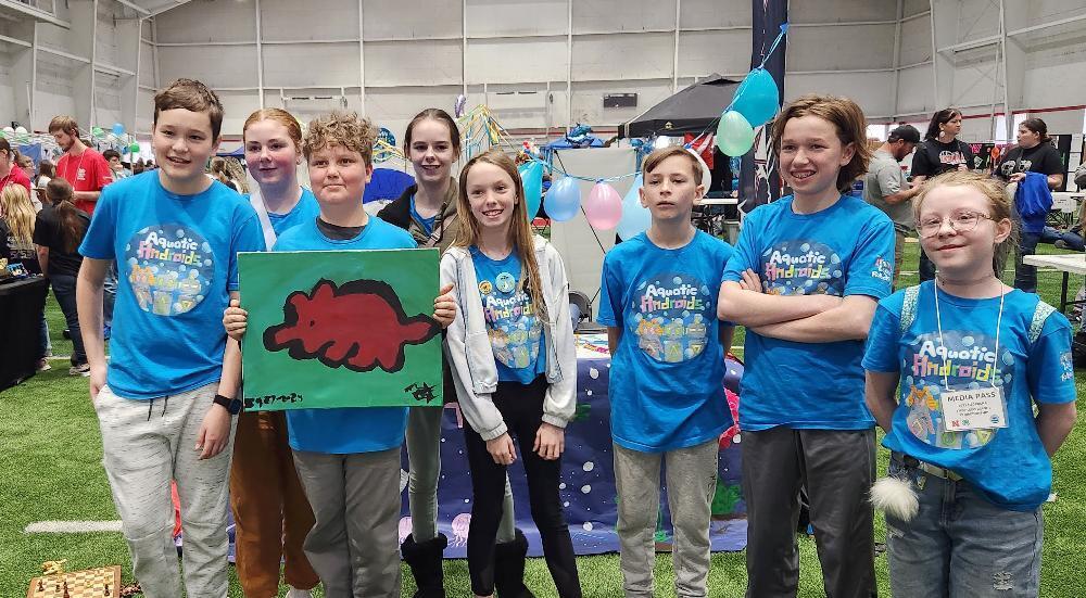 A group of children, wearing matching blue "Aquatic Androids" shirts, display a painted picture of a red dinosaur. They stand together in an indoor sports facility. One child wears a media pass.