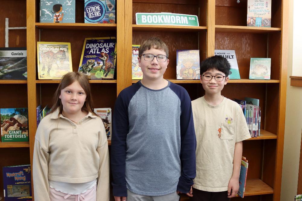 Three students stand in front of a bookshelf. They are positioned side by side, smiling at the camera. The shelves are filled with children's books on various topics.