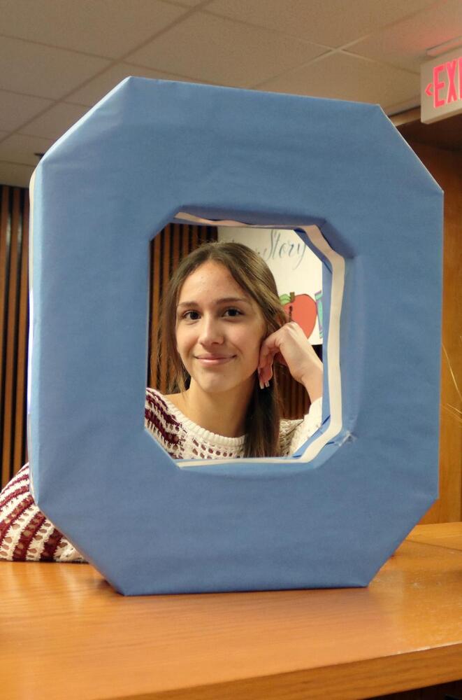 A smiling young woman peeks through a large blue O-shaped frame, resting her hand on her cheek. She wears a patterned sweater. The word "Story" and an apple graphic are visible behind her.
