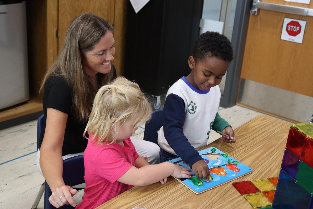 An adult paraeducator smiles as two preschool students play with an activity at a wooden table at Roper Elementary School.