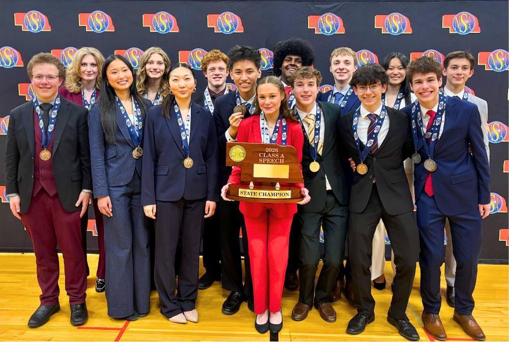 Sixteen smiling students, male and female, in suits and medals, proudly display their "State Champion Speech" trophy. A girl in a bright red suit holds the large wooden award centrally, celebrating their achievement.