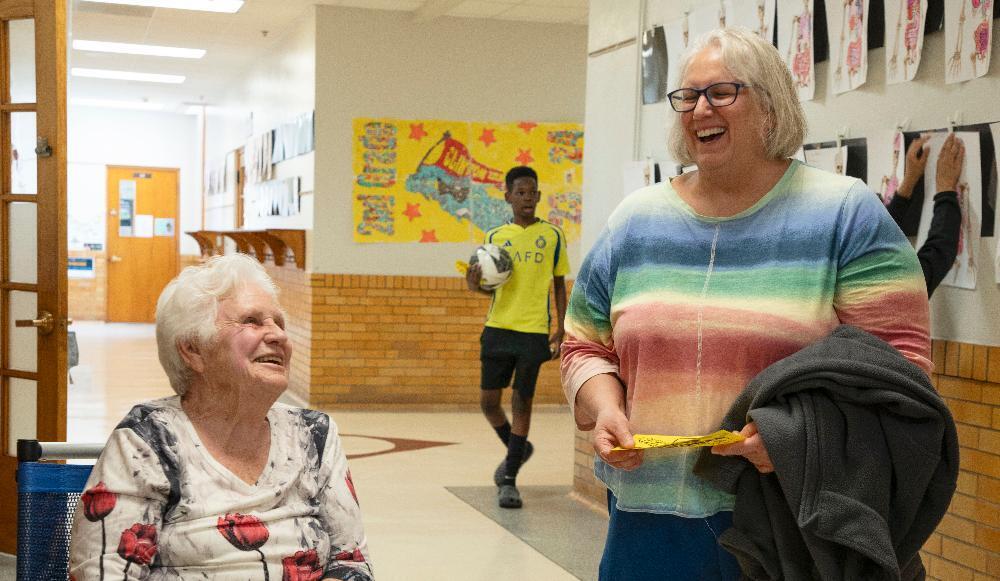 An elderly woman sits smiling in a bustling hallway. A laughing woman in a rainbow shirt holds papers. A boy walks by with a soccer ball, amidst colorful student artwork on the walls.